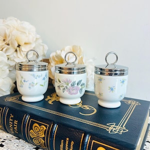 May include: Three white porcelain egg coddlers with silver-toned metal lids featuring circular ring handles. The coddlers are decorated with delicate floral patterns in blue, green, and pink, and are positioned atop a dark blue book titled Vanity Fair.