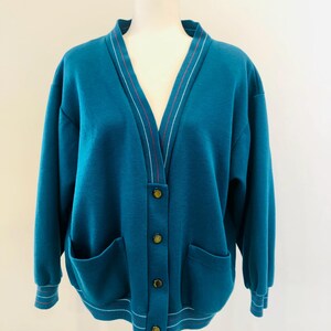 Peut inclure: Un cardigan bleu sarcelle à col en V, fermeture boutonnée et deux poches. Le cardigan est orné de rayures blanches et rouges autour du col et des poignets.