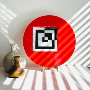 May include: A bright red decorative plate featuring a black and white geometric pattern at its centre. The plate is round and presented on a brass stand. Two decorative vases are in the background.