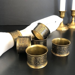 Peut inclure: Ensemble de six ronds de serviette anciens en laiton avec des motifs noirs gravés complexes. Les anneaux sont disposés autour d'une serviette en tissu blanche, avec deux bougeoirs en laiton et des bougies en arrière-plan. Parfait pour une table élégante.