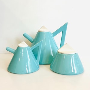 May include: A three-piece tea set featuring a teapot, a smaller creamer, and a sugar bowl. Each piece is finished in a glossy turquoise glaze with a distinct conical shape, a white triangular lid, and a geometric handle on the pouring vessels.