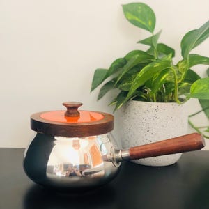 May include: A stainless steel saucepan with a wooden handle and lid. The lid has an orange centre and a wooden rim. The handle and lid are made of dark wood. The pot is reflective, and the background is blurred.