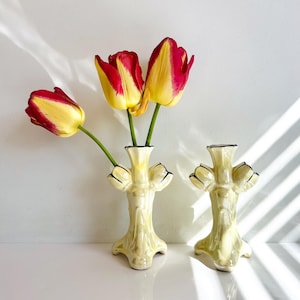 May include: Two pale yellow ceramic vases with black trim, each holding red and yellow tulips. The vases have a unique, stylized design with a flared base and three decorative elements near the top. The tulips add a pop of colour against the neutral background.