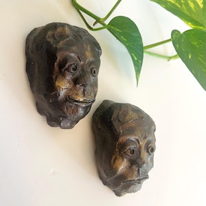 May include: Two dark brown, textured, hand-sculpted wall masks featuring human-like facial features with prominent eyes, noses, and mouths. The surfaces have a weathered, metallic bronze-toned finish with highlights on the forehead and cheeks.