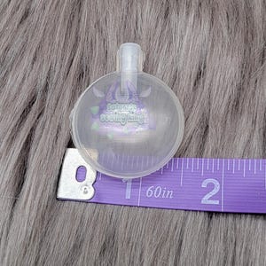 May include: A clear, round plastic object with a small spout, possibly a bottle stopper or similar item. The object is next to a purple measuring tape showing 2 inches. The object has the text "Sakura's Sewing Ring" printed on it.