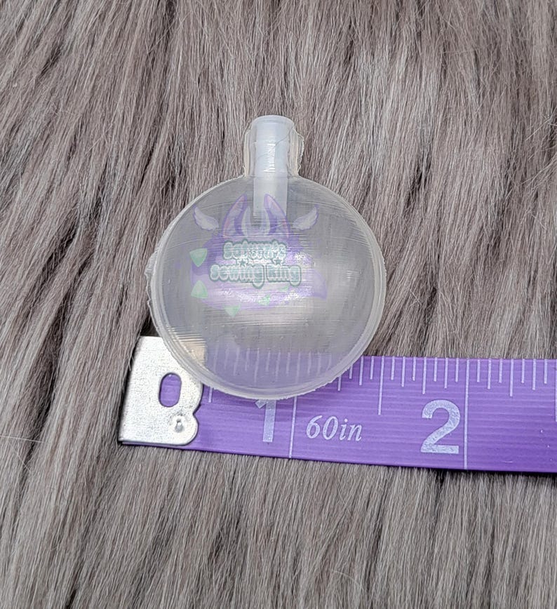 May include: A clear, round plastic object with a small spout, possibly a bottle stopper or similar item. The object is next to a purple measuring tape showing 2 inches. The object has the text "Sakura's Sewing Ring" printed on it.