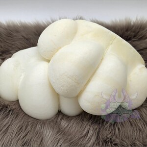 May include: A white, rounded, multi-lobed foam structure resembling a stylised animal paw. The object has a smooth, matte surface with several distinct, plump, cushion-like sections. A small purple logo with the text Saturn's Sewing King is visible.
