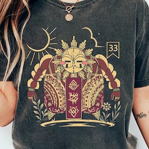 May include: Dark grey t-shirt with a maroon and gold illustration of a sun-like figure with a face, braids, and patterns. The number 33 is in a small box. The design includes a sun, clouds, and floral elements.
