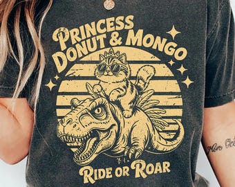 May include: Dark gray t-shirt with a graphic of a cat wearing sunglasses and a crown riding a dinosaur, with the text "Princess Donut & Mongo" and "Ride or Roar" in a retro style.