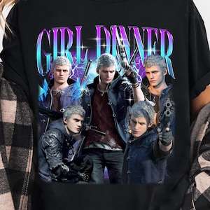 May include: Black t-shirt featuring a graphic with the text "GIRL DINNER" in a gradient blue and purple font. The graphic includes images of characters with silver hair and guns. The shirt is a casual style.