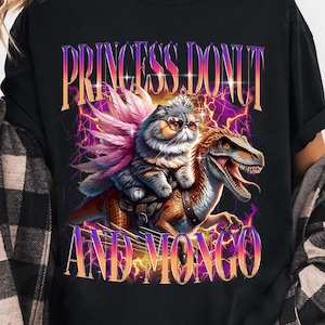May include: Black t-shirt featuring a graphic of a cat wearing sunglasses and riding a dinosaur, with the text "PRINCESS DONUT AND MONGO" in a purple and gold design. The cat has a pink feather-like accessory.
