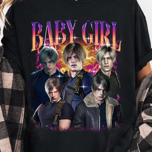 May include: Black t-shirt with a graphic print of five characters from the video game Resident Evil. The text "BABY GIRL" is printed in a retro style above the characters.