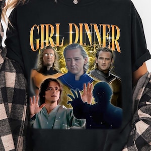 May include: Black t-shirt with a graphic featuring the text "GIRL DINNER" in gold lettering. The graphic includes images of several people. The person is wearing a black and white plaid shirt.