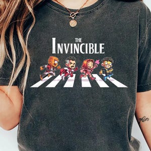 May include: A dark gray t-shirt with a graphic of the characters from the animated series "The Invincible" walking across a crosswalk. The text "The Invincible" is printed above the graphic.