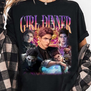 Può includere: Una t-shirt nera a maniche corte con la scritta GIRL DINNER in un gradiente viola e arancione. La grafica mostra cinque personaggi maschili in un'illustrazione stilizzata con uno sfondo caratterizzato da fulmini gialli e viola.