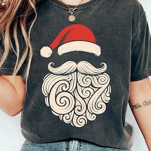 May include: Dark gray t-shirt featuring a Santa Claus design. The design includes a red Santa hat, a white mustache, and a beard with the number 67. The shirt is made of a soft material.