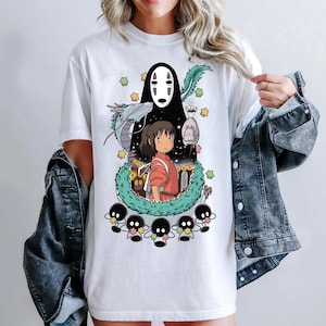 Może przedstawiać: Biała koszulka z nadrukiem graficznym postaci z filmu animowanego Spirited Away. Grafika przedstawia czarno-białego ducha, młodą dziewczynę z czerwonymi włosami i zieloną, podobną do smoka istotę. Grafika jest otoczona małymi, czarno-białymi stworzeniami.