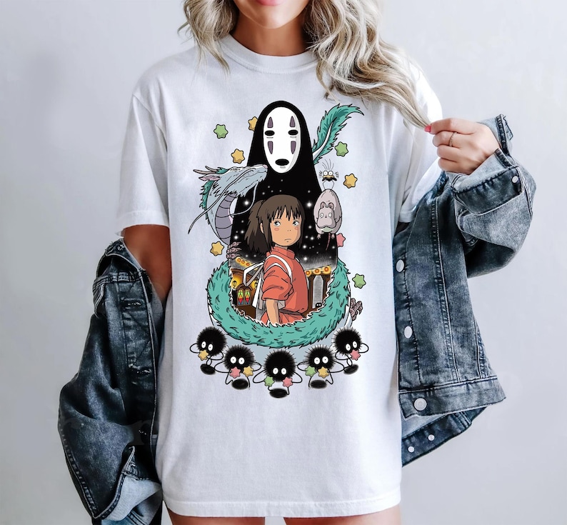 Może przedstawiać: Biała koszulka z nadrukiem graficznym postaci z filmu animowanego Spirited Away. Grafika przedstawia czarno-białego ducha, młodą dziewczynę z czerwonymi włosami i zieloną, podobną do smoka istotę. Grafika jest otoczona małymi, czarno-białymi stworzeniami.