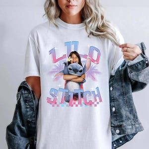 Pode incluir: Camiseta branca com um gr&aacute;fico de Stitch de Lilo & Stitch abra&ccedil;ando uma menina. O design inclui as palavras "Lilo" e "Stitch" em uma fonte gradiente rosa e azul. Palmeiras est&atilde;o no fundo.