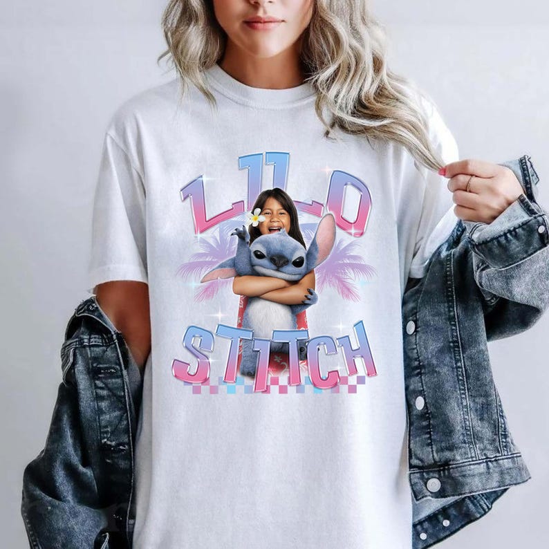 Pode incluir: Camiseta branca com um gr&aacute;fico de Stitch de Lilo & Stitch abra&ccedil;ando uma menina. O design inclui as palavras "Lilo" e "Stitch" em uma fonte gradiente rosa e azul. Palmeiras est&atilde;o no fundo.