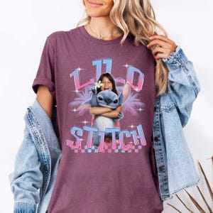 Pode incluir: Camiseta bord&ocirc; com um desenho de Stitch de Lilo & Stitch abra&ccedil;ando uma menina. O design inclui o texto "11.0 Stitch" em uma fonte gradiente, palmeiras e estrelas.