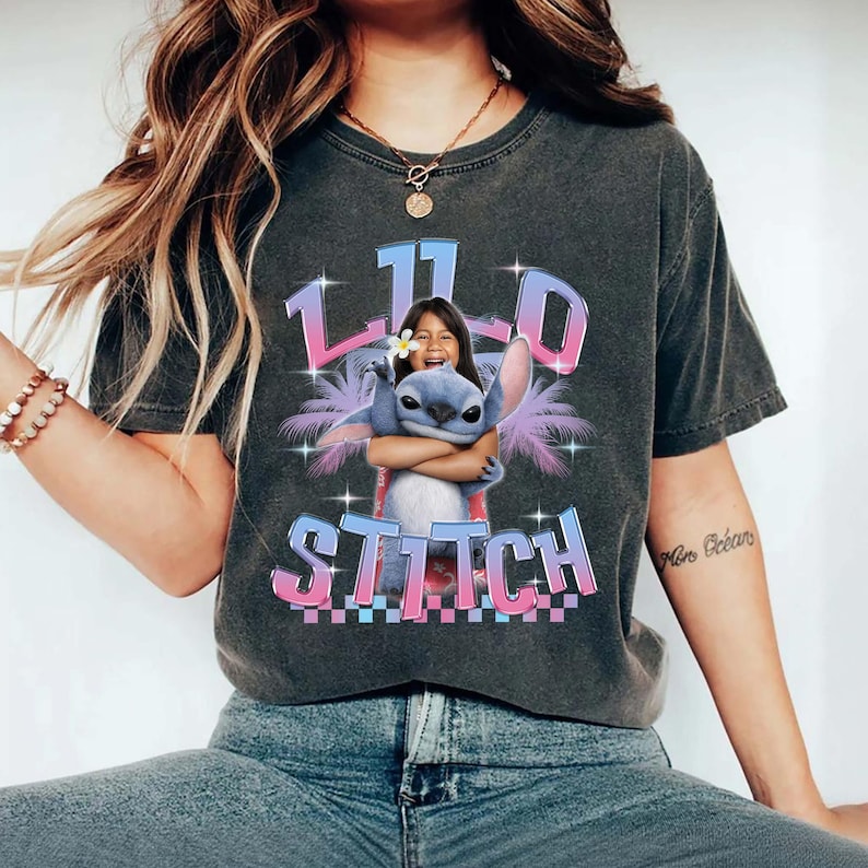 Pode incluir: Camiseta cinza escuro com um gr&aacute;fico de Stitch abra&ccedil;ando uma menina e o texto "11th Stitch" em gradiente rosa e azul. O design inclui palmeiras e estrelas, criando um tema tropical.