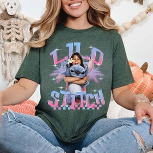 Pode incluir: T-shirt verde escuro com um gr&aacute;fico de uma menina abra&ccedil;ando o personagem Stitch. O design inclui o texto "110 Stitch" em uma fonte colorida e estilizada. A camiseta tem um corte folgado.