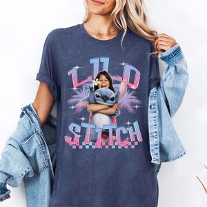 Pode incluir: Uma camiseta azul marinho com um gr&aacute;fico de uma menina abra&ccedil;ando o personagem Stitch do filme Lilo & Stitch. O gr&aacute;fico inclui o texto "11" e "Stitch" em um design tropical colorido. A camiseta tem mangas curtas.