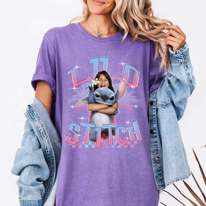 Pode incluir: Uma camiseta roxa com um gr&aacute;fico de uma menina abra&ccedil;ando o personagem Stitch. O gr&aacute;fico inclui o texto "11" e "Stitch" em uma fonte colorida e estilizada. A camiseta tem mangas curtas e um corte folgado.