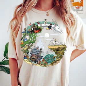 K&ouml;nnte beinhalten: Cremefarbenes T-Shirt mit einem kreisf&ouml;rmigen Grafikdesign mit animierten Charakteren und Szenen. Das Design umfasst einen wei&szlig;en Drachen, ein M&auml;dchen auf einem Besen und verschiedene Geb&auml;ude und Kreaturen in einer farbenfrohen, detaillierten Illustration.