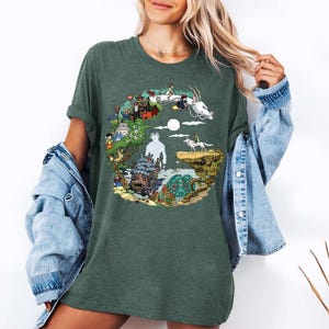 K&ouml;nnte beinhalten: Gr&uuml;nes T-Shirt mit einem farbenfrohen Grafikdesign, das Charaktere und Szenen aus einem Zeichentrickfilm zeigt. Das Design umfasst einen wei&szlig;en Drachen, ein wolfs&auml;hnliches Wesen sowie verschiedene Geb&auml;ude und Figuren. Das Shirt hat kurze &Auml;rmel und eine lockere Passform.