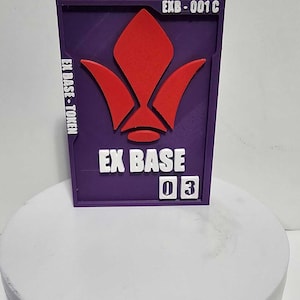 K&ouml;nnte beinhalten: Ein rechteckiger violetter Token mit einem erhabenen roten Emblem, wei&szlig;em Text mit der Aufschrift EX BASE, EX BASE-TOKEN und EXB-001C sowie zwei wei&szlig;en quadratischen Pl&auml;ttchen mit der Zahl 03. Der Token ist flach und vertikal ausgerichtet.