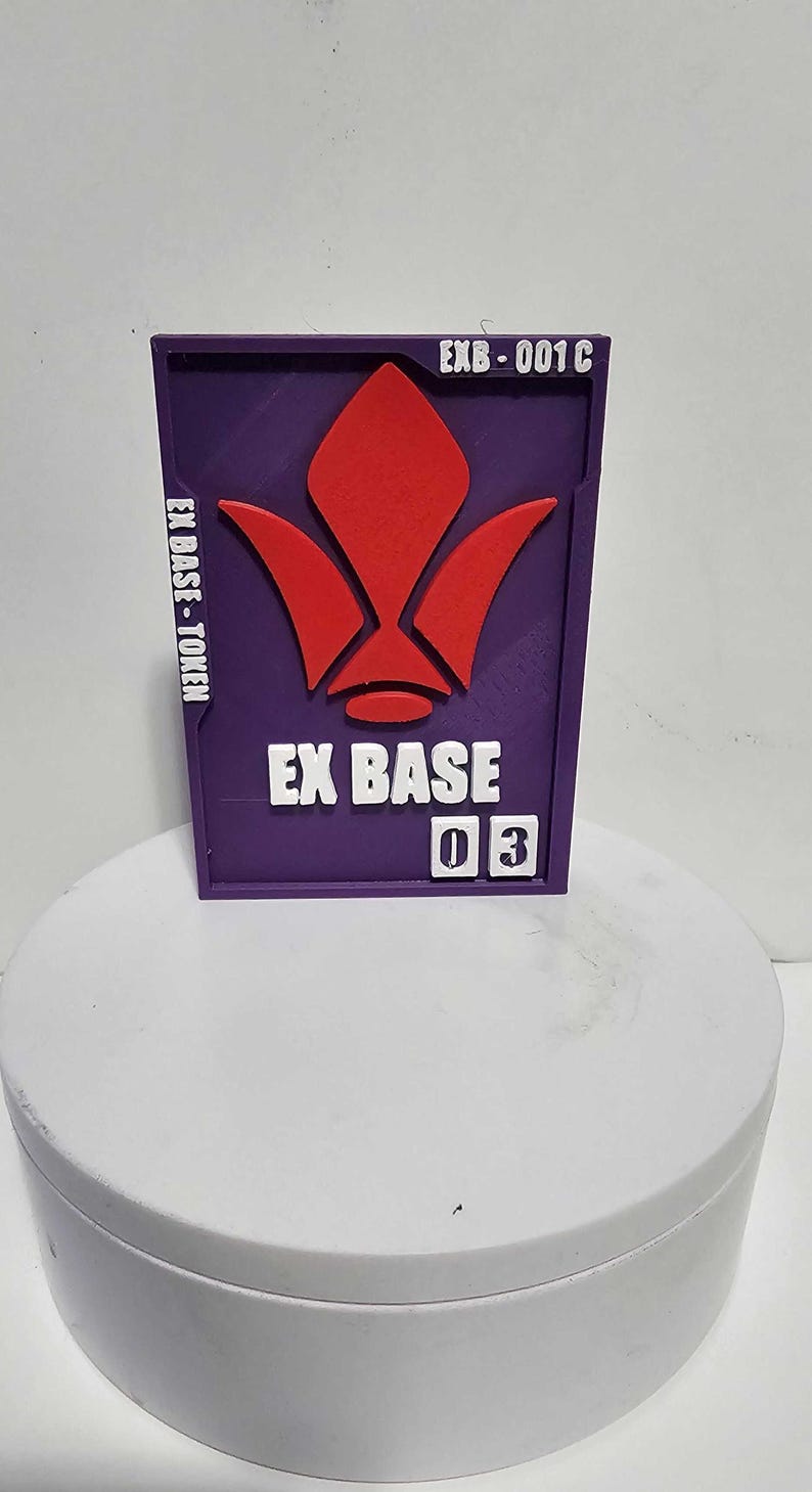 K&ouml;nnte beinhalten: Ein rechteckiger violetter Token mit einem erhabenen roten Emblem, wei&szlig;em Text mit der Aufschrift EX BASE, EX BASE-TOKEN und EXB-001C sowie zwei wei&szlig;en quadratischen Pl&auml;ttchen mit der Zahl 03. Der Token ist flach und vertikal ausgerichtet.