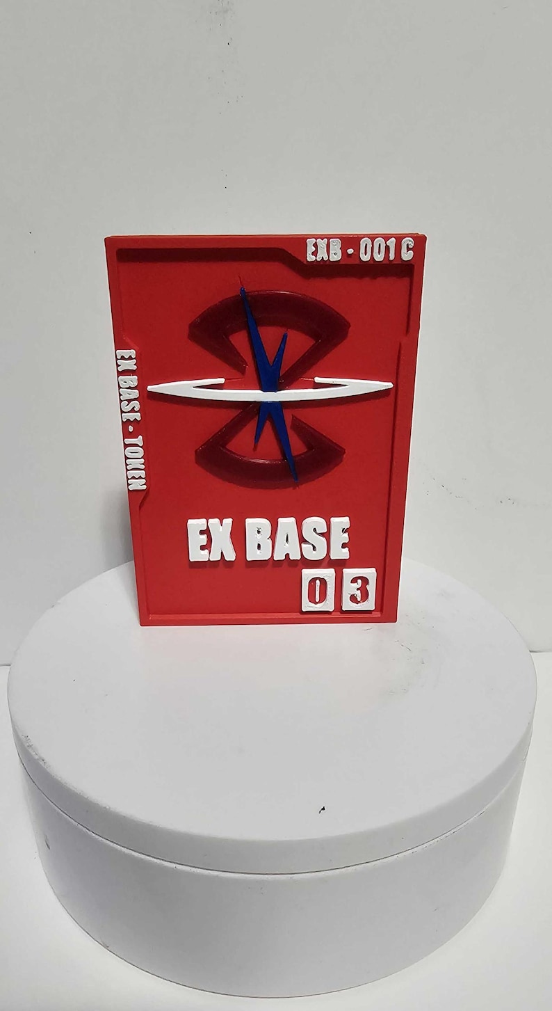 K&ouml;nnte beinhalten: Ein rechteckiger roter Token mit dem Text EXB-001C oben rechts, EX BASE-TOKEN auf der linken Seite und EX BASE 03 unten. In der Mitte befindet sich ein erhabenes dunkelrotes kreisf&ouml;rmiges Emblem mit einer blauen vertikalen und einer wei&szlig;en horizontalen Linie.