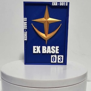 K&ouml;nnte beinhalten: Eine rechteckige blaue Plakette mit einem goldfarbenen Stern- und Halbmondemblem. Der Text EX BASE - TOKEN ist links vertikal aufgedruckt, EXB-001 C oben rechts und EX BASE 03 unten in wei&szlig;en, erhabenen Buchstaben.