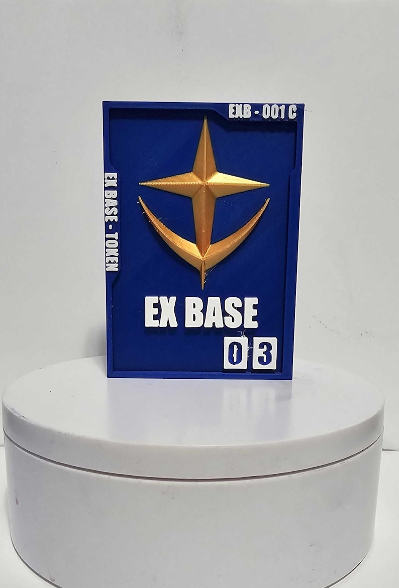 K&ouml;nnte beinhalten: Eine rechteckige blaue Plakette mit einem goldfarbenen Stern- und Halbmondemblem. Der Text EX BASE - TOKEN ist links vertikal aufgedruckt, EXB-001 C oben rechts und EX BASE 03 unten in wei&szlig;en, erhabenen Buchstaben.
