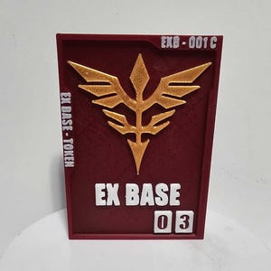 K&ouml;nnte beinhalten: Eine rechteckige kastanienbraune Plakette mit einem erhabenen goldenen Emblem in Form eines stilisierten vogelartigen Wappens. Der Text EX BASE - TOKEN steht links, EXB-001C oben rechts und die Zahl 03 in wei&szlig;en K&auml;stchen unten rechts.