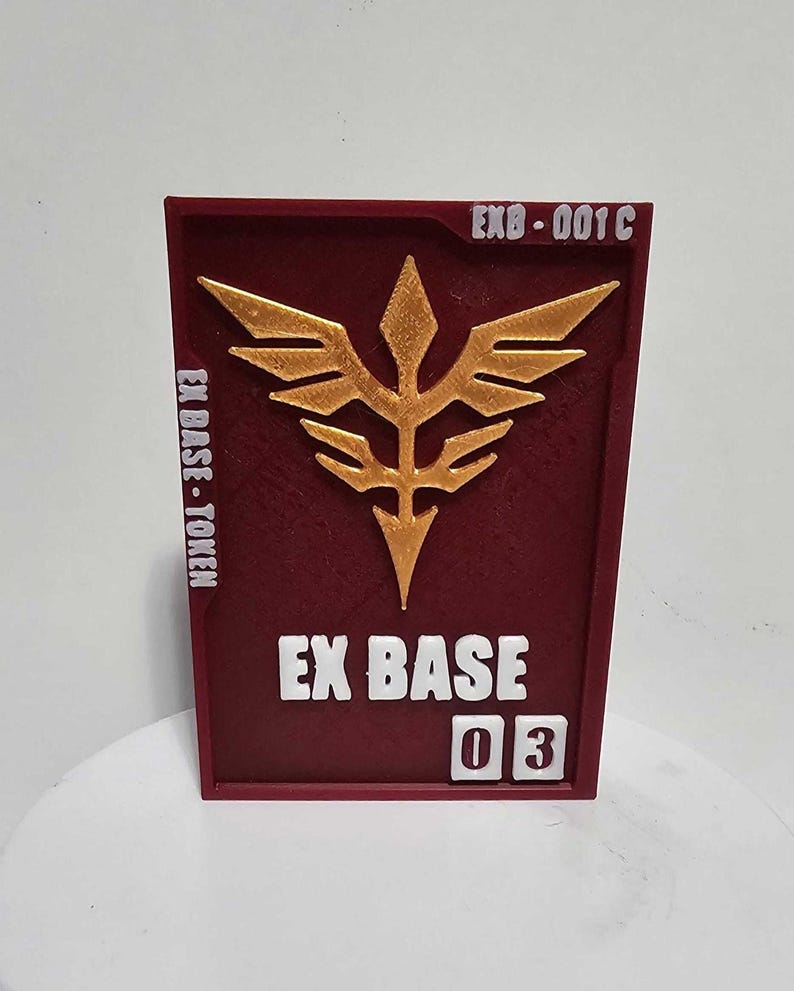 K&ouml;nnte beinhalten: Eine rechteckige kastanienbraune Plakette mit einem erhabenen goldenen Emblem in Form eines stilisierten vogelartigen Wappens. Der Text EX BASE - TOKEN steht links, EXB-001C oben rechts und die Zahl 03 in wei&szlig;en K&auml;stchen unten rechts.