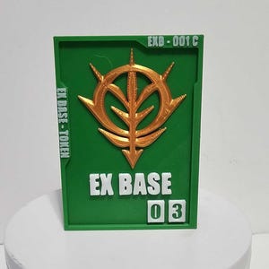 K&ouml;nnte beinhalten: Eine rechteckige gr&uuml;ne Plakette mit einem erhabenen goldenen Emblem in der Mitte. Der Text EXB-001 C steht oben rechts, EX BASE-TOKEN befindet sich auf der linken Seite und EX BASE 03 ist unten in Wei&szlig; mit der Zahl 03 in wei&szlig;en Quadraten.