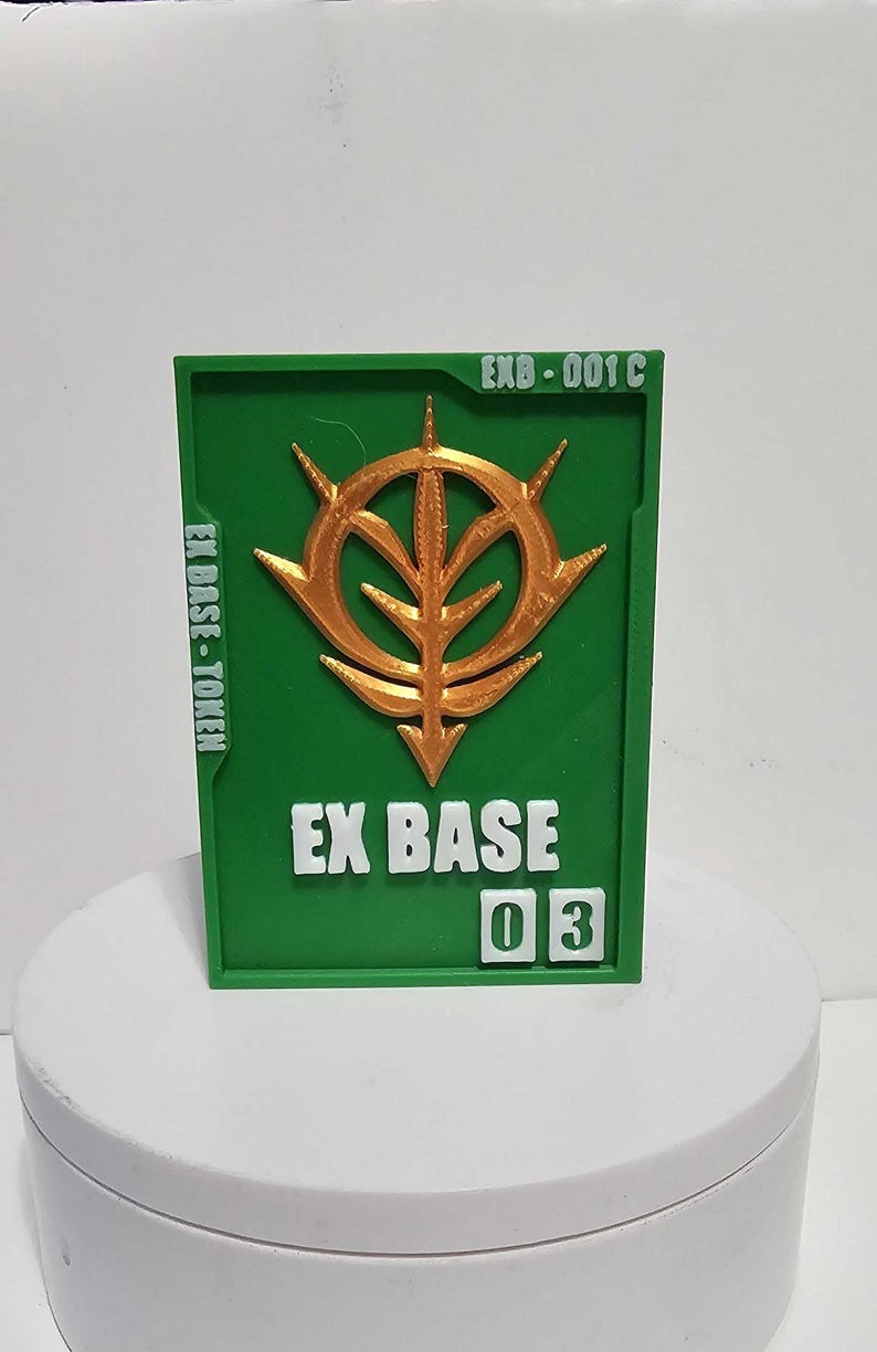 K&ouml;nnte beinhalten: Eine rechteckige gr&uuml;ne Plakette mit einem erhabenen goldenen Emblem in der Mitte. Der Text EXB-001 C steht oben rechts, EX BASE-TOKEN befindet sich auf der linken Seite und EX BASE 03 ist unten in Wei&szlig; mit der Zahl 03 in wei&szlig;en Quadraten.