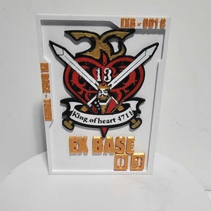 K&ouml;nnte beinhalten: Eine rechteckige wei&szlig;e Plakette mit einem zentralen roten Herz-Emblem, das eine K&ouml;nigsfigur, gekreuzte Schwerter und die Zahl 13 zeigt. Der Text lautet EXB-0016, EX BASE-TOKEN und EX BASE 03 in goldener Schrift. Enth&auml;lt den Text King of heart 4711.