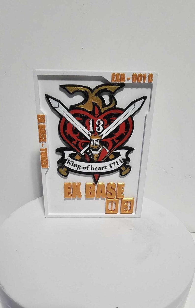 K&ouml;nnte beinhalten: Eine rechteckige wei&szlig;e Plakette mit einem zentralen roten Herz-Emblem, das eine K&ouml;nigsfigur, gekreuzte Schwerter und die Zahl 13 zeigt. Der Text lautet EXB-0016, EX BASE-TOKEN und EX BASE 03 in goldener Schrift. Enth&auml;lt den Text King of heart 4711.
