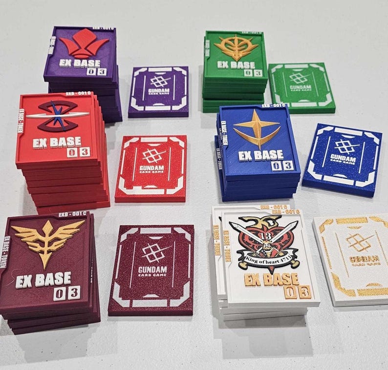 May include: A collection of rectangular plastic game tokens in various colors including red, blue, green, purple, and white. Each token features the text EX BASE 03, a unique emblem, and the Gundam Card Game logo on the reverse side of the stacks.