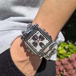 May include: A silver-toned, square-faced wristwatch with a white dial and black and red accents. The watch features multiple subdials and the brand name "Oakley". The watch is worn on a silver link bracelet.