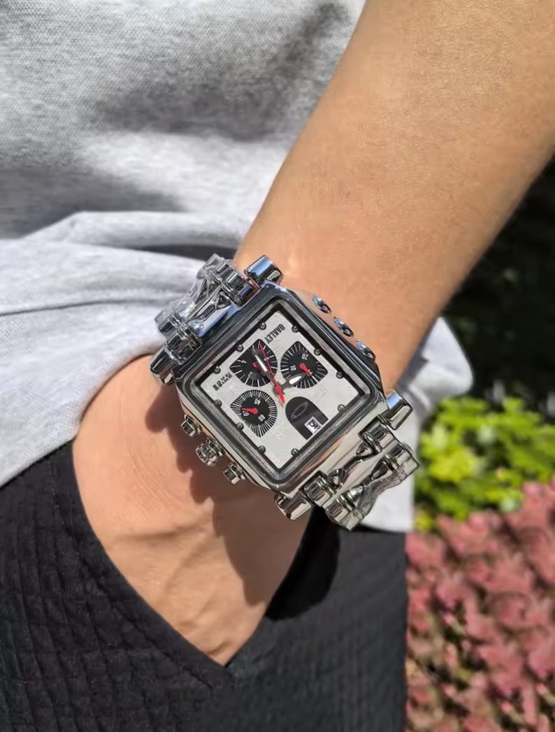 May include: A silver-toned, square-faced wristwatch with a white dial and black and red accents. The watch features multiple subdials and the brand name "Oakley". The watch is worn on a silver link bracelet.