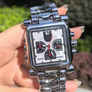 May include: A silver-toned Oakley watch with a square face and a matching metal band. The watch face is white with black sub-dials and red hands. The watch has the brand name "Oakley" printed on the face.