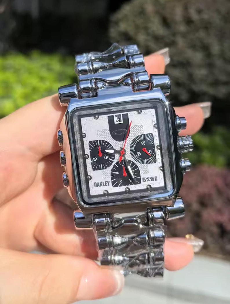 May include: A silver-toned Oakley watch with a square face and a matching metal band. The watch face is white with black sub-dials and red hands. The watch has the brand name "Oakley" printed on the face.