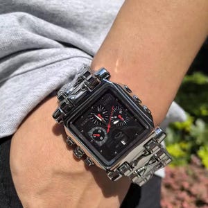 May include: A silver-toned wristwatch with a square black face and a matching silver band. The watch features multiple sub-dials with red accents and a textured design. The watch is on a wrist.