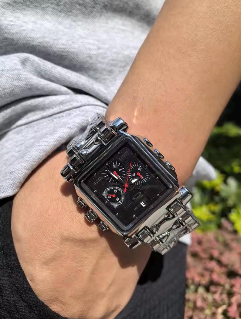 May include: A silver-toned wristwatch with a square black face and a matching silver band. The watch features multiple sub-dials with red accents and a textured design. The watch is on a wrist.