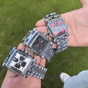 Puede incluir: Tres relojes de pulsera cuadrados con correas de eslabones de metal plateado. Las esferas de los relojes son negras, blancas y rojas, con múltiples esferas y la marca "BAILEE". Los relojes se sostienen sobre un fondo verde.